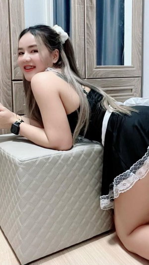 Asian woman in black maid dress leaning on pouf indoors.