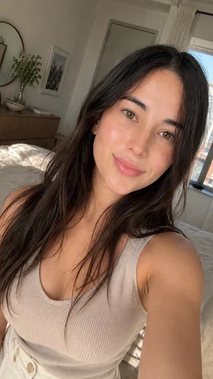 Woman with long dark hair in a beige tank top, smiling in a bedroom selfie.