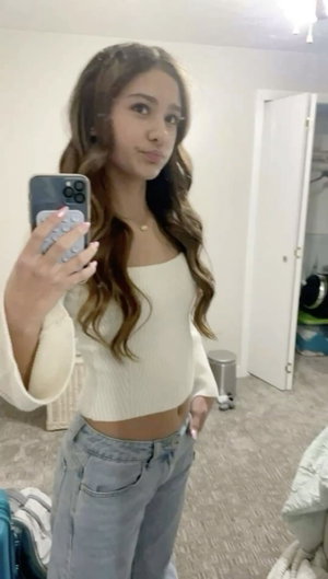 Woman with long wavy hair in cream crop top and jeans taking mirror selfie in bedroom.