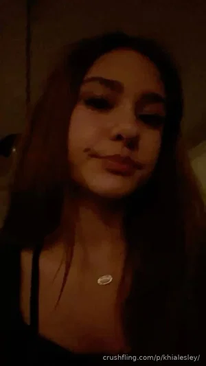 Woman with long dark hair wearing black top and necklace, indoors in dim light.