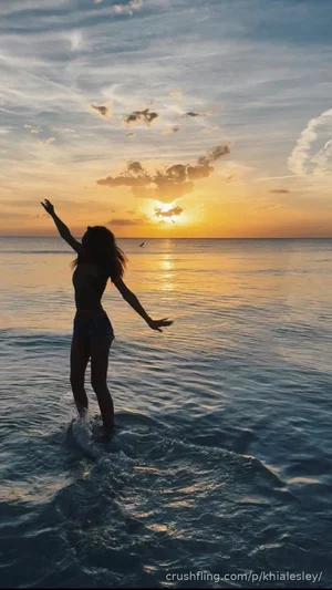 Woman in bikini silhouette standing in ocean at sunset, arms raised.