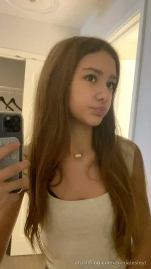 Woman with long brown hair in a white top taking a mirror selfie in a bedroom.
