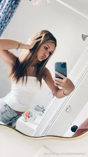 Woman with long hair in white top and denim shorts takes mirror selfie in bathroom.