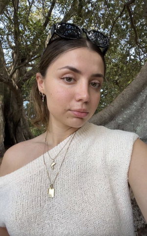 Woman with brown hair in white sweater outdoors near trees.