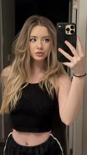 Woman with blonde hair in black tank top and sweatpants, indoor selfie.