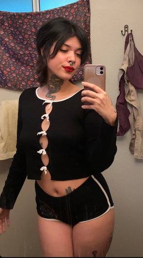 Woman with dark hair and tattoos taking mirror selfie indoors.