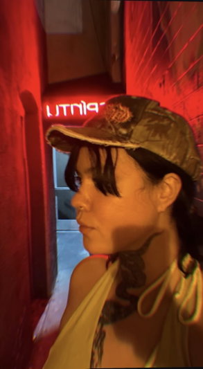 Woman in yellow top and cap with neck tattoos in red-lit alley.