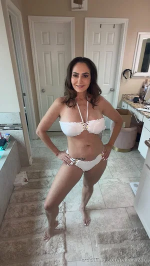 Woman in white bikini posing in bathroom, hands on hips, smiling.