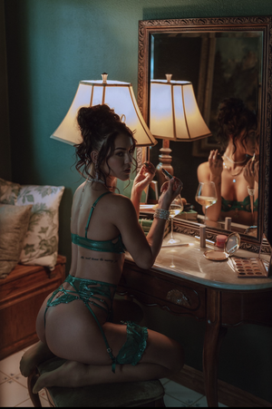 Woman in green lingerie applying makeup at vanity mirror in dimly lit bedroom.