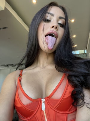 Woman with long dark hair in red sheer top, sticking tongue out indoors.
