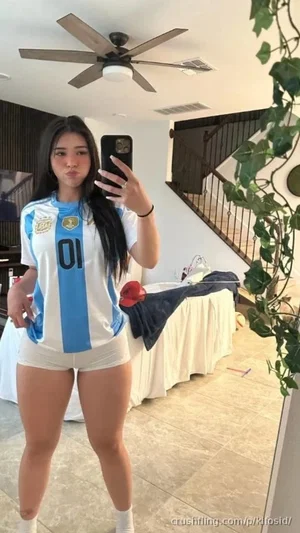 Woman in Argentina soccer jersey and white shorts taking mirror selfie indoors.