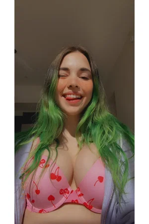 Woman with green hair wearing pink cherry-print bra and light jacket, smiling indoors.