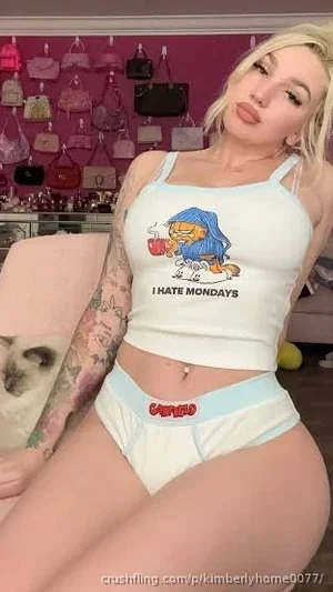 Woman with blonde hair and tattoos wears a white crop top and light blue underwear, sitting in a