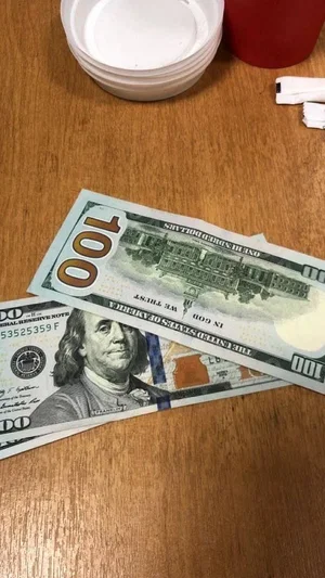 Two hundred-dollar bills on a wooden table with plastic cups and a red cup nearby.