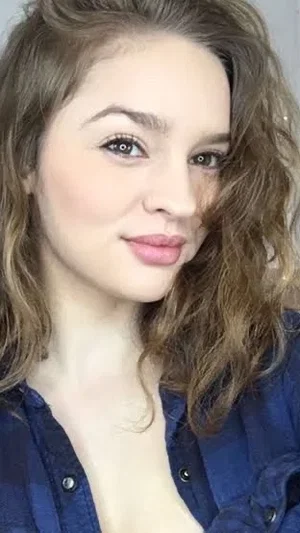 Woman with wavy brown hair in a blue button-up shirt, close-up selfie indoors.