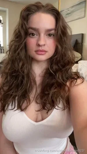 Woman with curly brown hair in white tank top, indoors, facing camera.