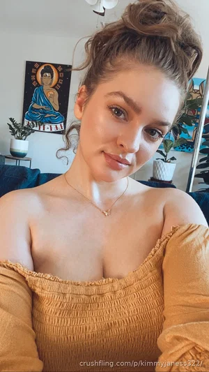 Woman with brown hair in a bun wearing an off-shoulder top, indoors with plants and artwork in the