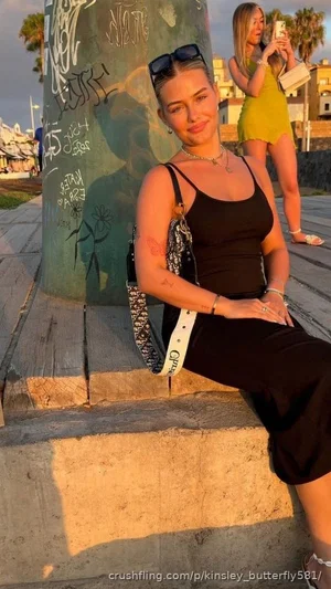 Woman in black tank top and skirt sits on concrete steps outdoors, sunglasses on head, bag on