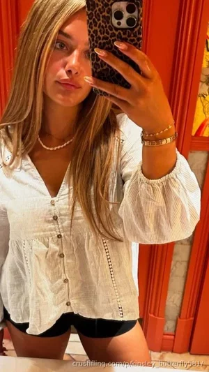 Woman with long blonde hair in white blouse and black shorts taking mirror selfie indoors.
