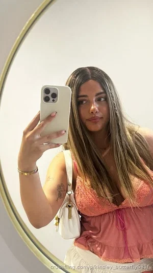 Woman with long hair in pink lace top and white skirt taking mirror selfie indoors.