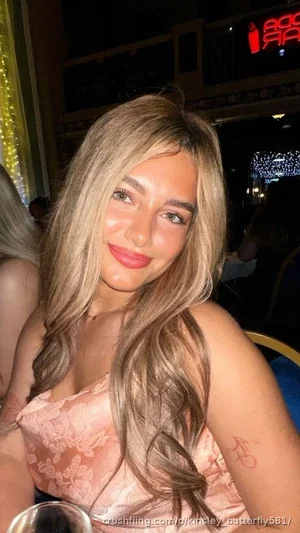 Woman with long blonde hair in a pink lace top, smiling in a dimly lit indoor setting.