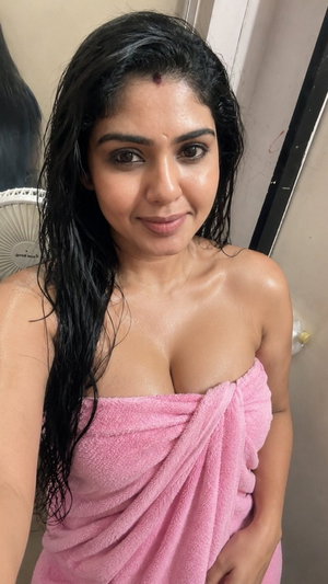 Woman with wet dark hair wrapped in pink towel, standing indoors near fan.