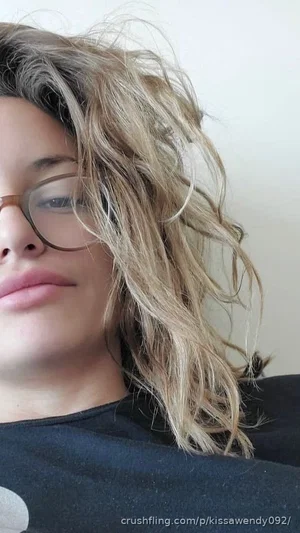 Woman with wavy blonde hair and glasses, wearing dark top, close-up selfie indoors.