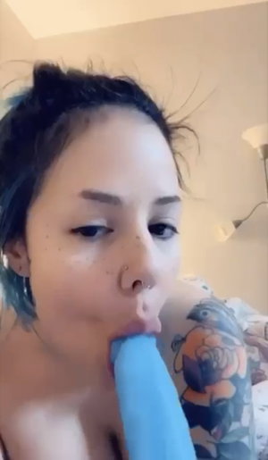 Dark-haired woman with tattoos holds blue object in mouth indoors.