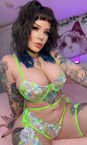 Woman with tattoos in floral lingerie posing in a bedroom with purple lighting.