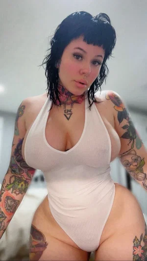Woman with tattoos in white one-piece swimsuit, indoor setting.