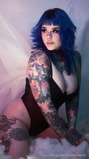 Woman with blue hair and tattoos in black bodysuit, posing on white fluffy surface.
