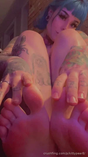 Woman with blue hair and tattoos on legs, sitting indoors, feet close to camera.