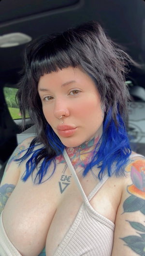 Tattooed woman with blue hair in low-cut top inside car.