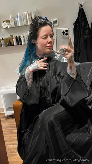 Woman with blue hair and tattoos in salon, wearing black cape, taking mirror selfie.