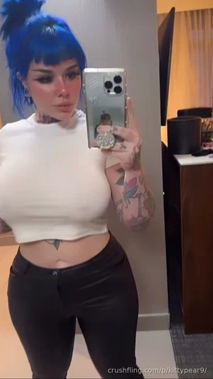 Woman with blue hair and tattoos wears white crop top and black pants, taking mirror selfie indoors.