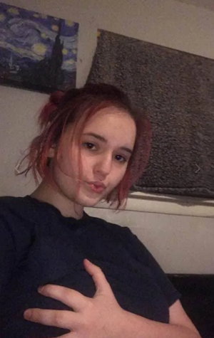 Red-haired woman in black t-shirt posing indoors with hand on chest.