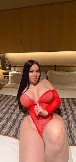 Curvy woman in red mesh bodysuit on bed in bedroom.
