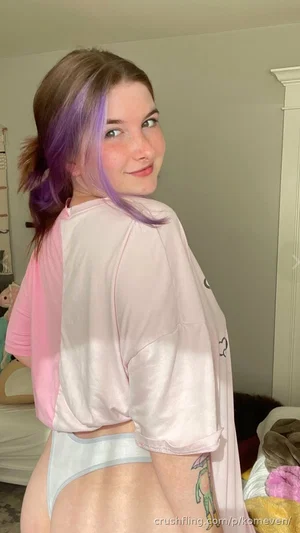 Woman with purple-tipped hair in a pink crop top and light blue underwear, standing in a bedroom.