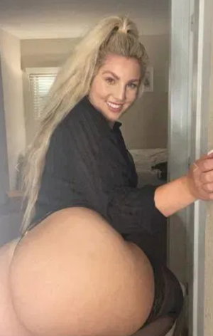 Blonde woman in black shirt, bare buttocks, posing in doorway, indoor setting.