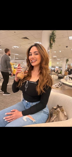 Woman with long brown hair in black top and ripped jeans, holding drink in store.