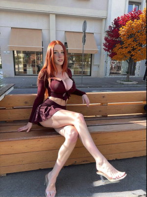 Woman with red hair in maroon outfit sits on wooden bench outdoors.