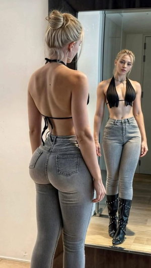 Woman in brown top and red leggings sits on stool, taking mirror selfie indoors.