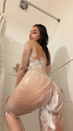 Woman with dark hair covered in soap, posing in shower, back to camera.