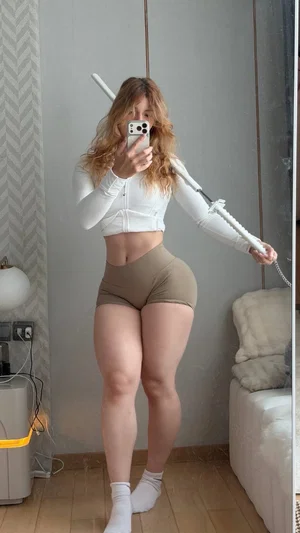Woman in white crop top and beige shorts holding a prop sword, taking mirror selfie in bedroom.