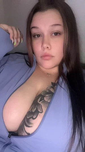 Woman with long dark hair and chest tattoo wearing low-cut blue top, indoor selfie.