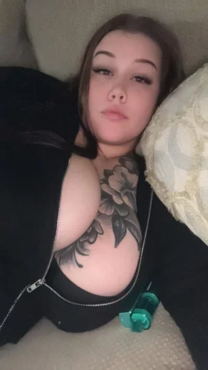 Woman with floral chest tattoo lying on couch, wearing unzipped black top.