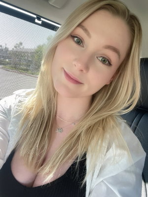 Blonde woman in black top and white jacket sitting in car, smiling at camera.