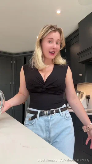 Woman with blonde hair in black top and jeans standing in kitchen.