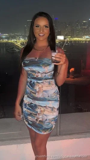 Woman in form-fitting printed dress holding drink, standing on balcony with city lights behind.