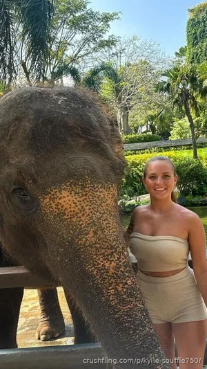 Woman in beige crop top and shorts posing with elephant outdoors in green setting.